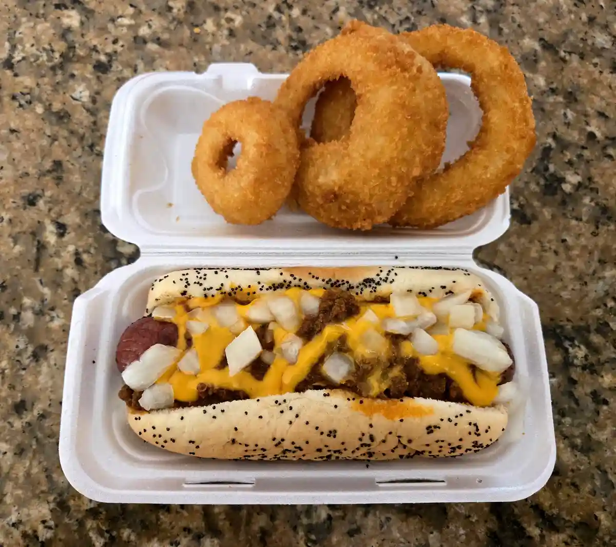 A white styrofoam takeout container holds a hot dog in a poppy seed bun topped with chili, melted yellow cheese, and diced…