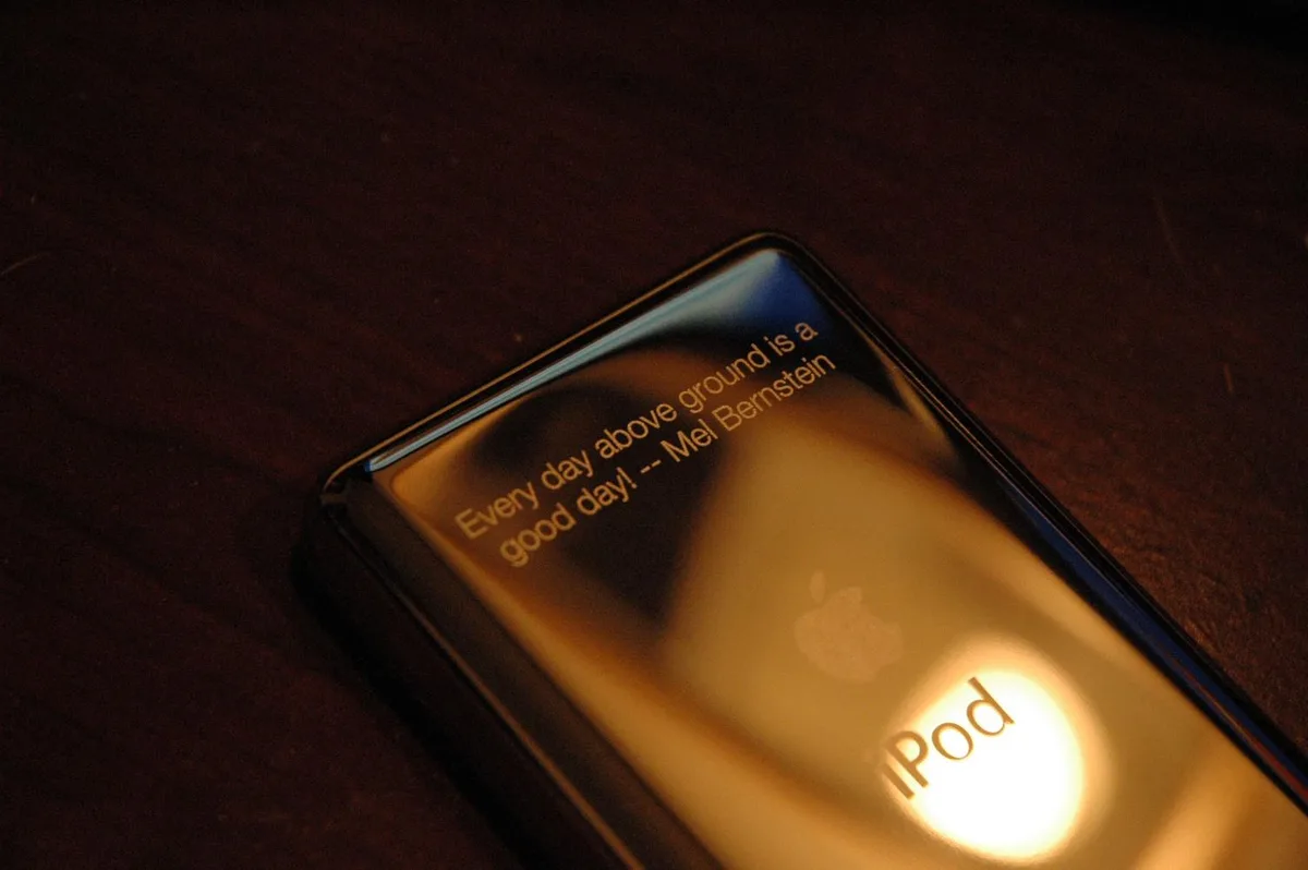 A close-up photo of a black Apple iPod resting on a dark wooden surface, its glossy back reflecting warm light. The text…