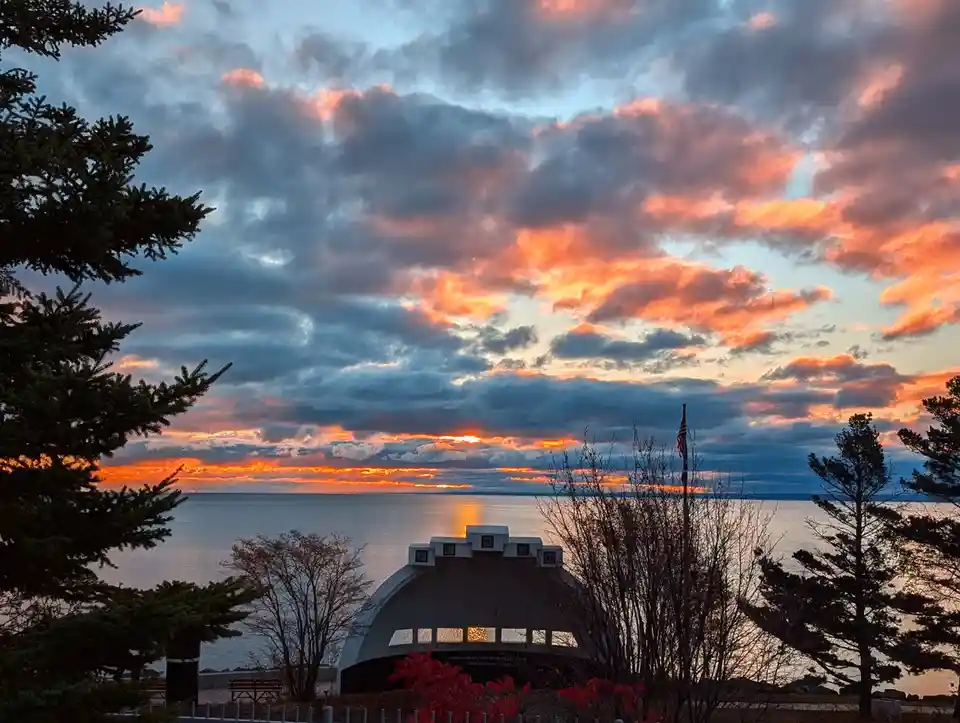A scenic sunset over a large body of water with dramatic orange and blue clouds. In the foreground, a white, arched memorial…
