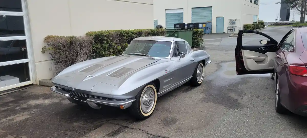 A silver 1960s Chevrolet Corvette with whitewall tires is parked next to a modern maroon sedan with its door open. The…