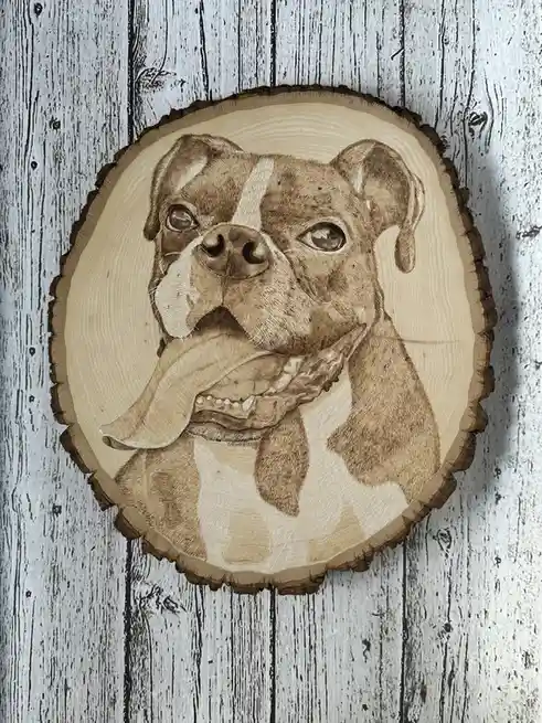 A detailed wood-burned portrait of a boxer dog with a white stripe on its face, mounted on a rustic wooden slice against a…