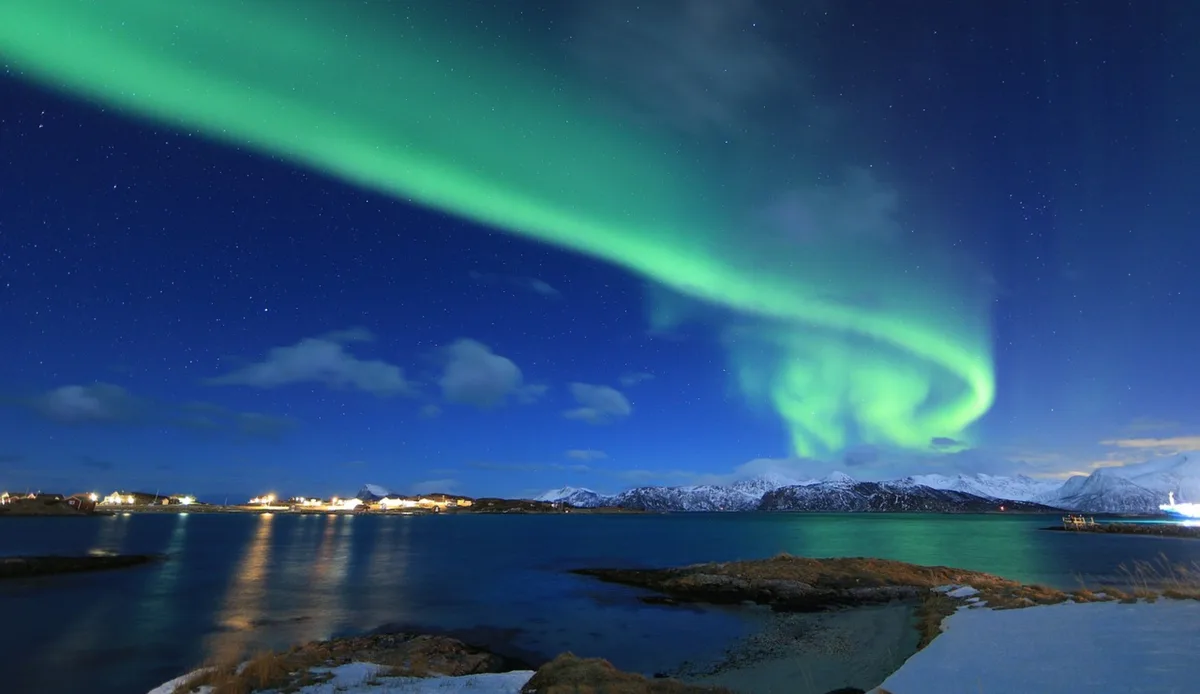 A stunning nighttime landscape photograph shows vibrant green aurora borealis dancing across a starry sky above a snowy…