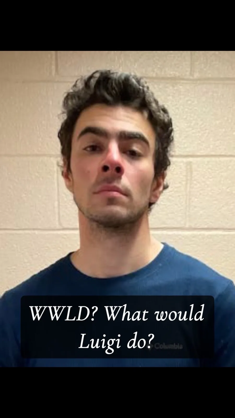 A man with dark curly hair and a blue t-shirt stands against a beige cinder block wall, looking directly at the camera with…