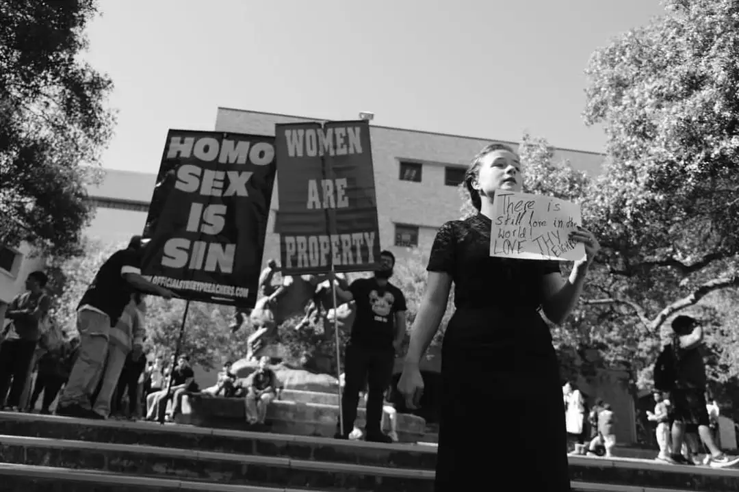 A black-and-white photo shows a woman in a dark dress holding a handwritten sign that reads 'There is Still love in the…