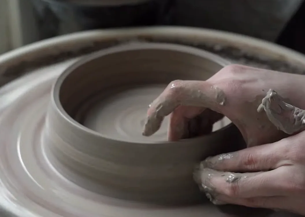 A close-up shot shows a person's hands covered in wet clay, shaping a spinning pottery wheel into a bowl. The clay is grey…