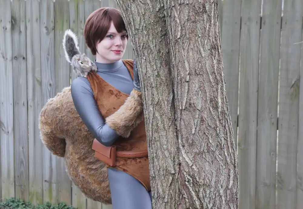 A person in a brown, furry squirrel costume with a grey turtleneck and leather belt is peeking from behind a tree trunk…