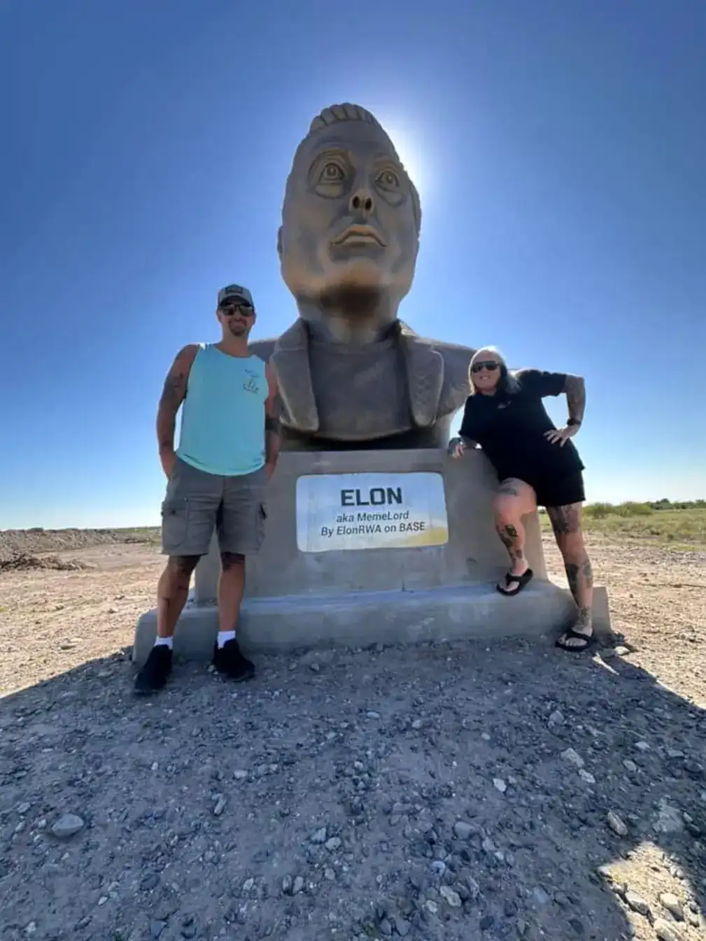 Two people pose beside a large outdoor bust of Elon Musk, which has a plaque identifying him as 'ELON aka MemeLord By…