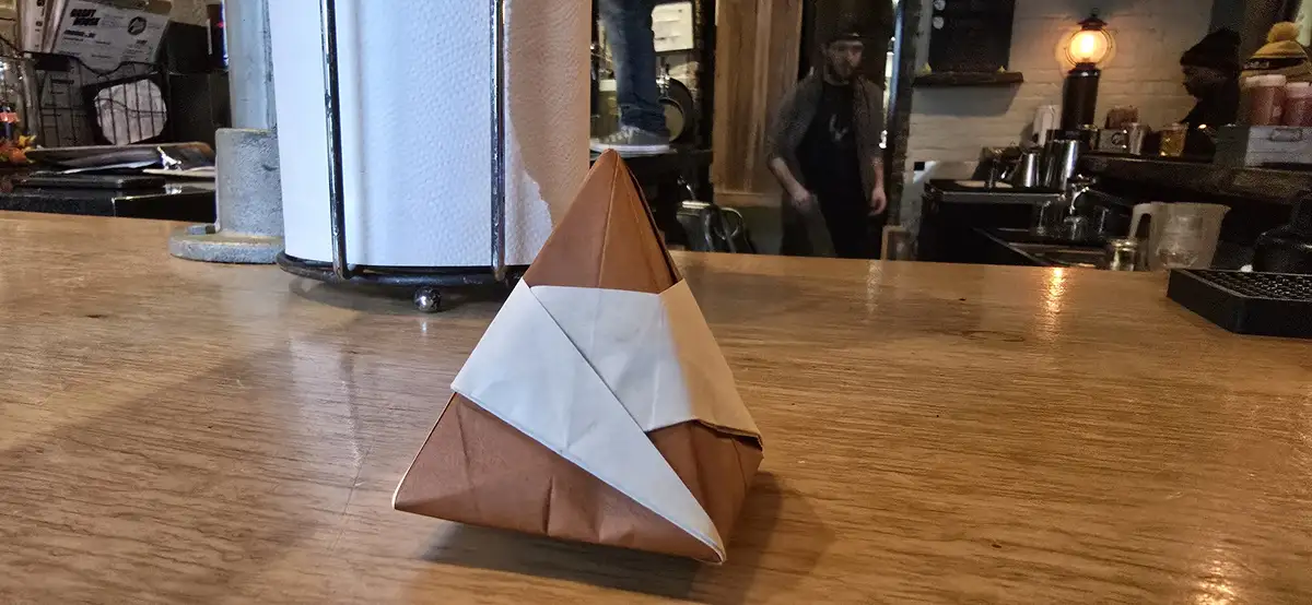 A paper origami fox sits on a wooden counter in a cafe, crafted from brown and white paper with a triangular shape…