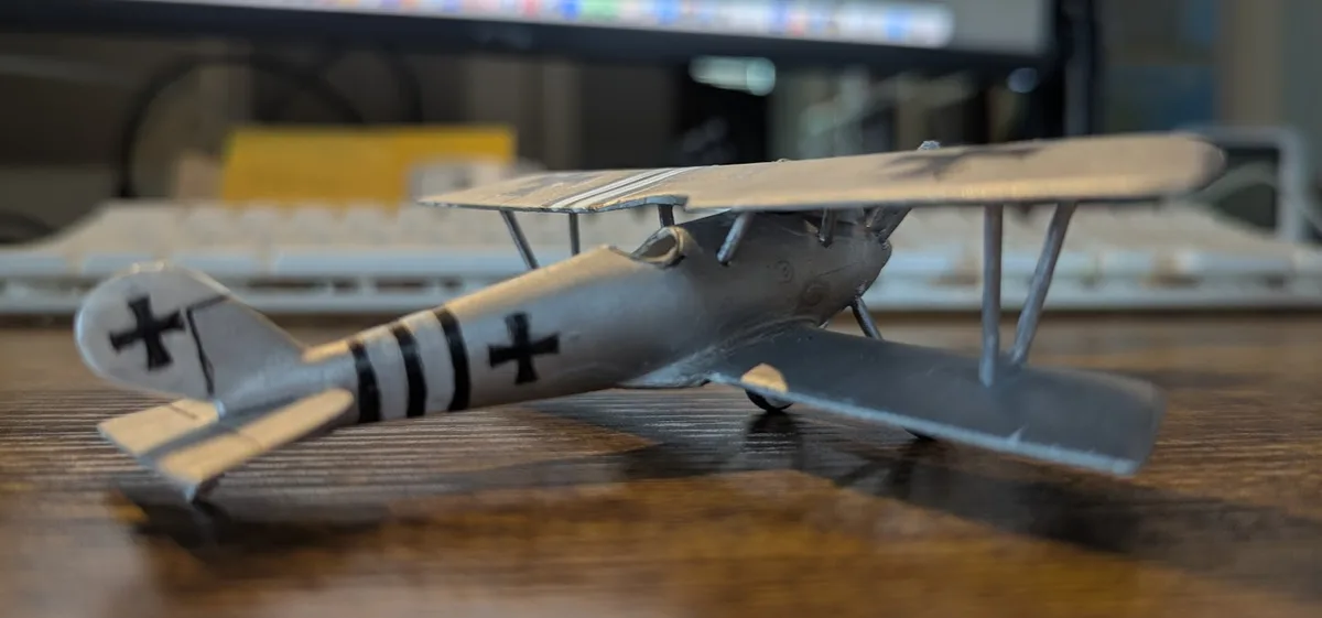 A silver model of a World War I-era biplane with black crosses and stripes sits on a wooden desk, with a blurred computer…