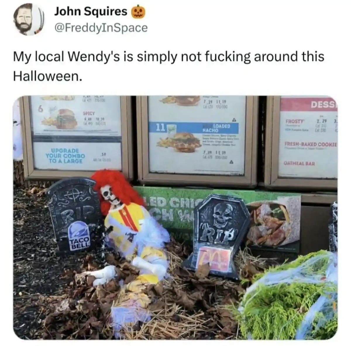 An outdoor Wendy's restaurant window displays its menu, with a Halloween-themed scene staged in front: a skeleton dressed as…