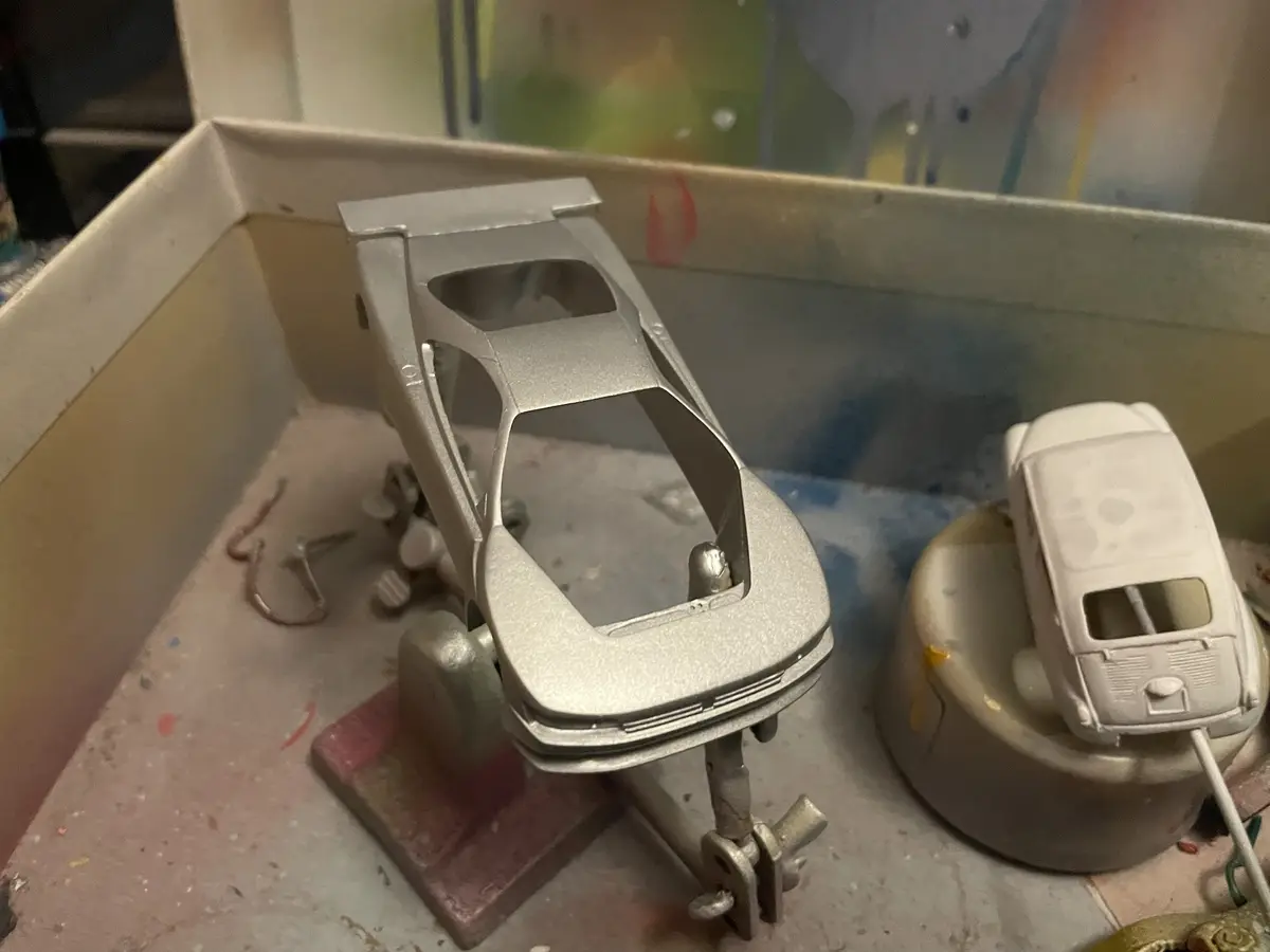 A close-up photo shows two model cars in a messy workshop. One is a silver, unpainted car body held in a vise, and the other…