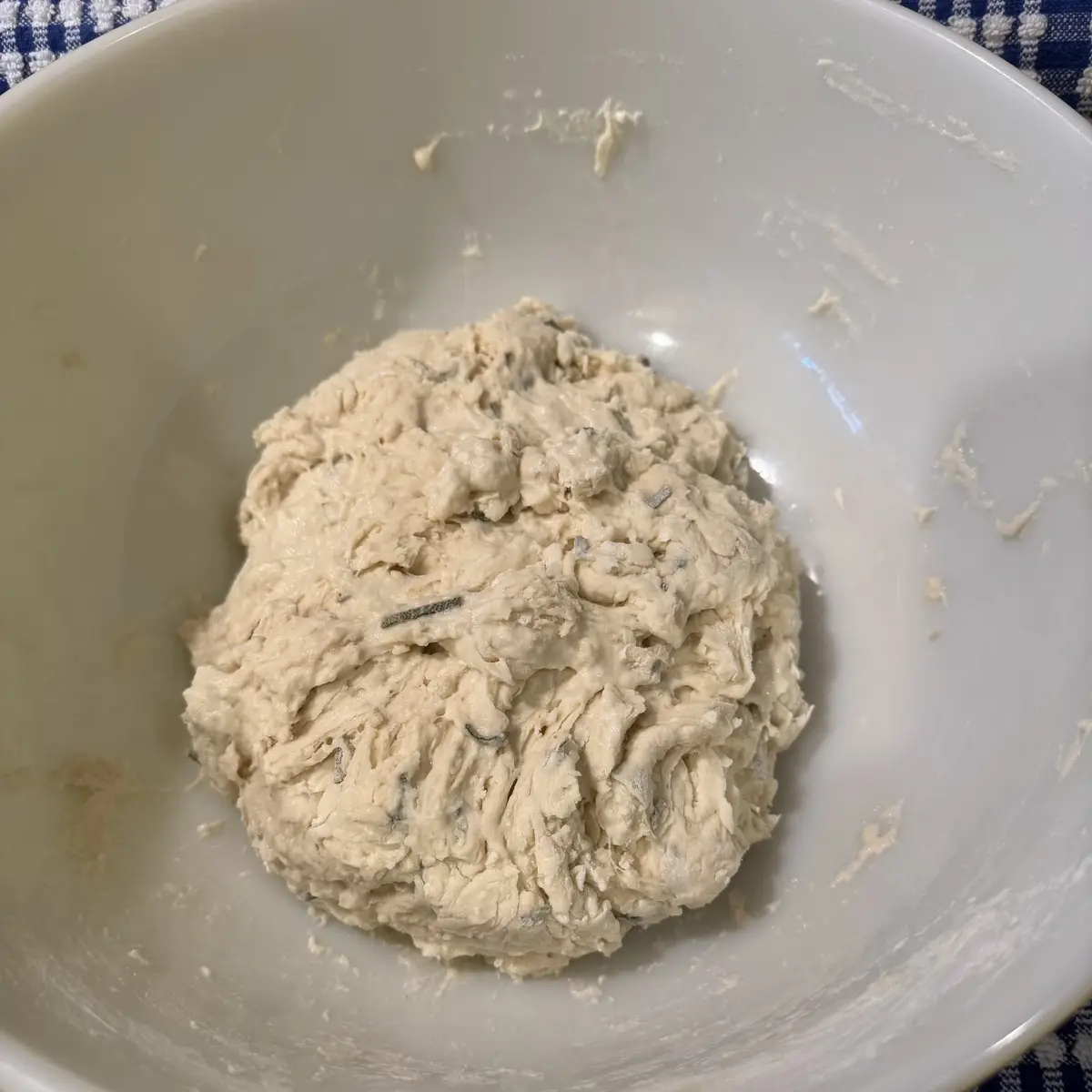 A close-up photo shows a lumpy, pale dough ball in a white mixing bowl, with visible green herb pieces embedded in it. The…