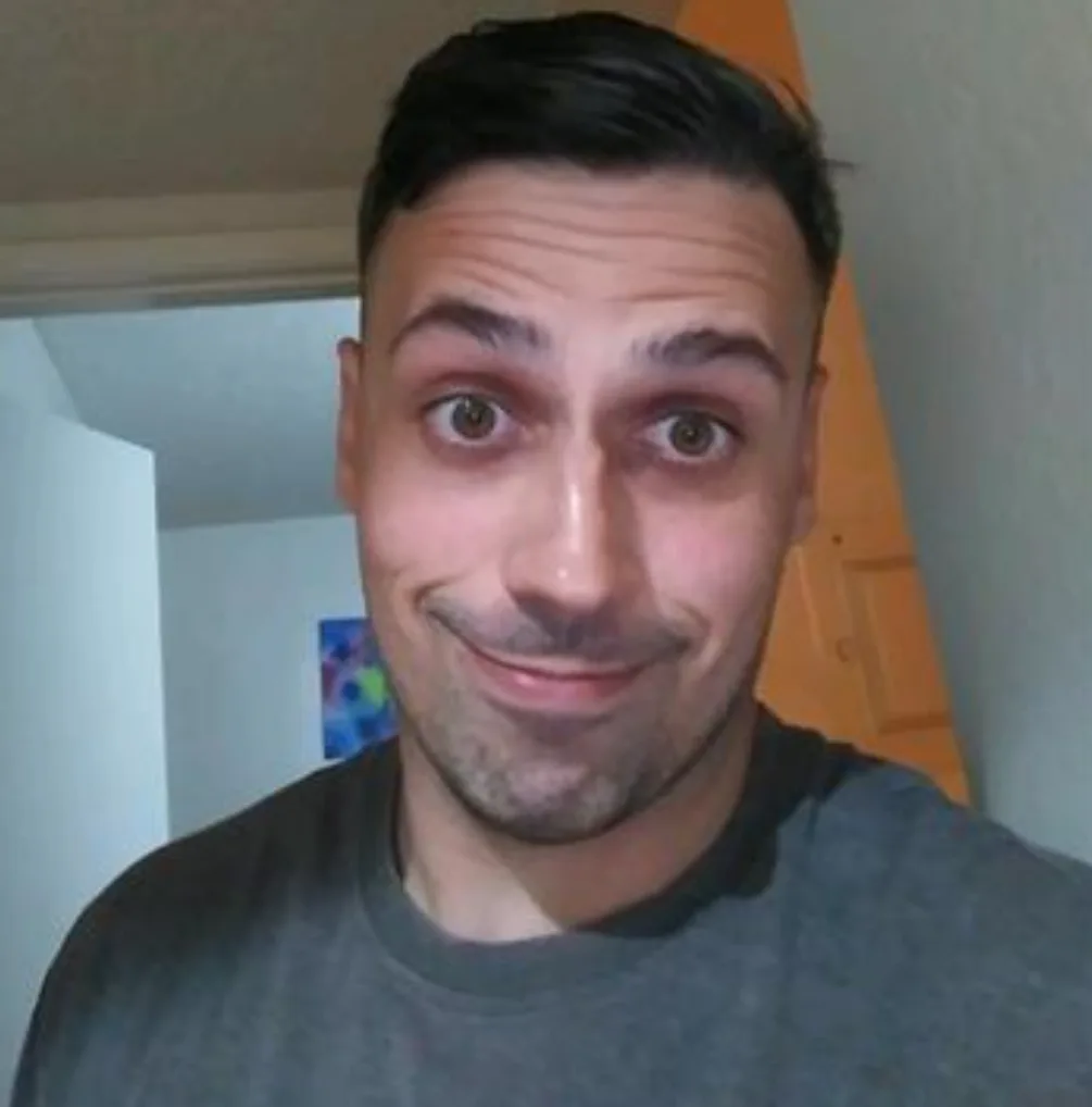 A man with dark hair and a gray t-shirt stares directly at the camera with wide, startled eyes and a forced, awkward smile…
