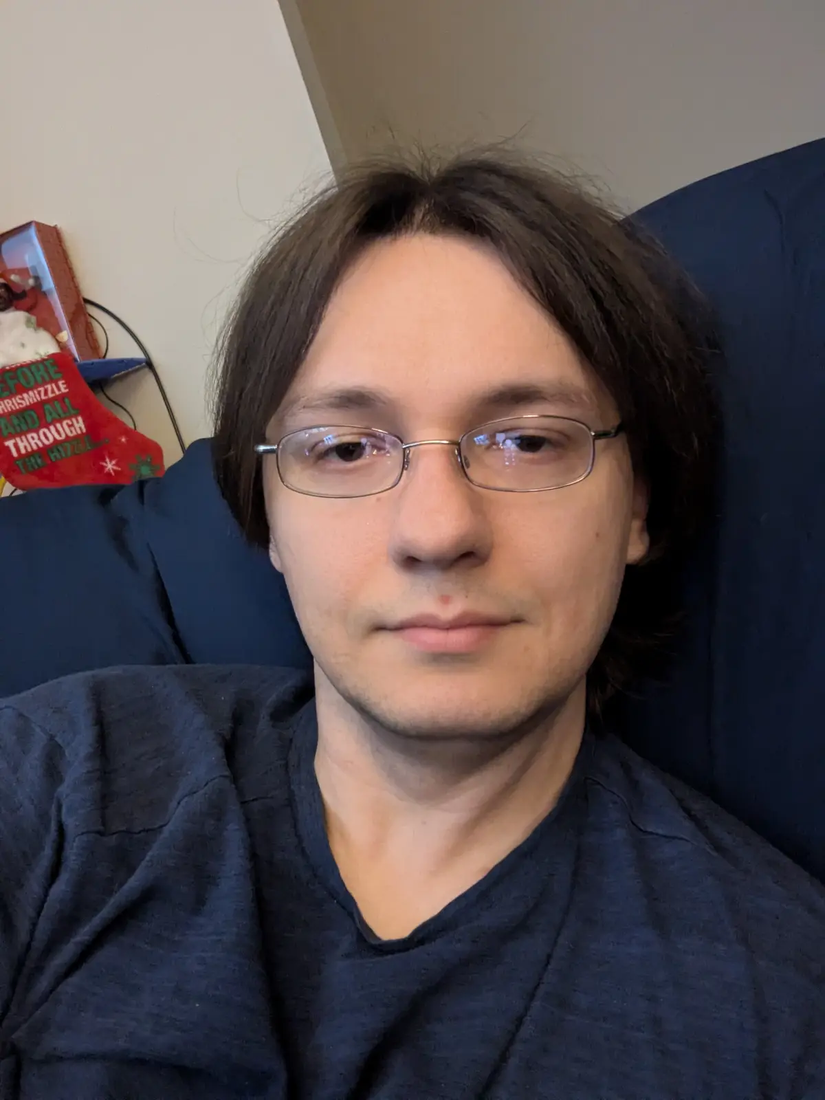 A man with glasses and dark hair sits in a blue chair wearing a dark blue t-shirt, looking directly at the camera with a…