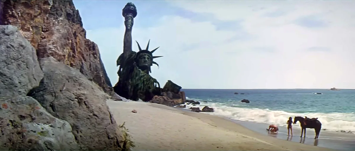 A weathered, partially submerged Statue of Liberty stands on a rocky beach next to the ocean, while two people and a horse…