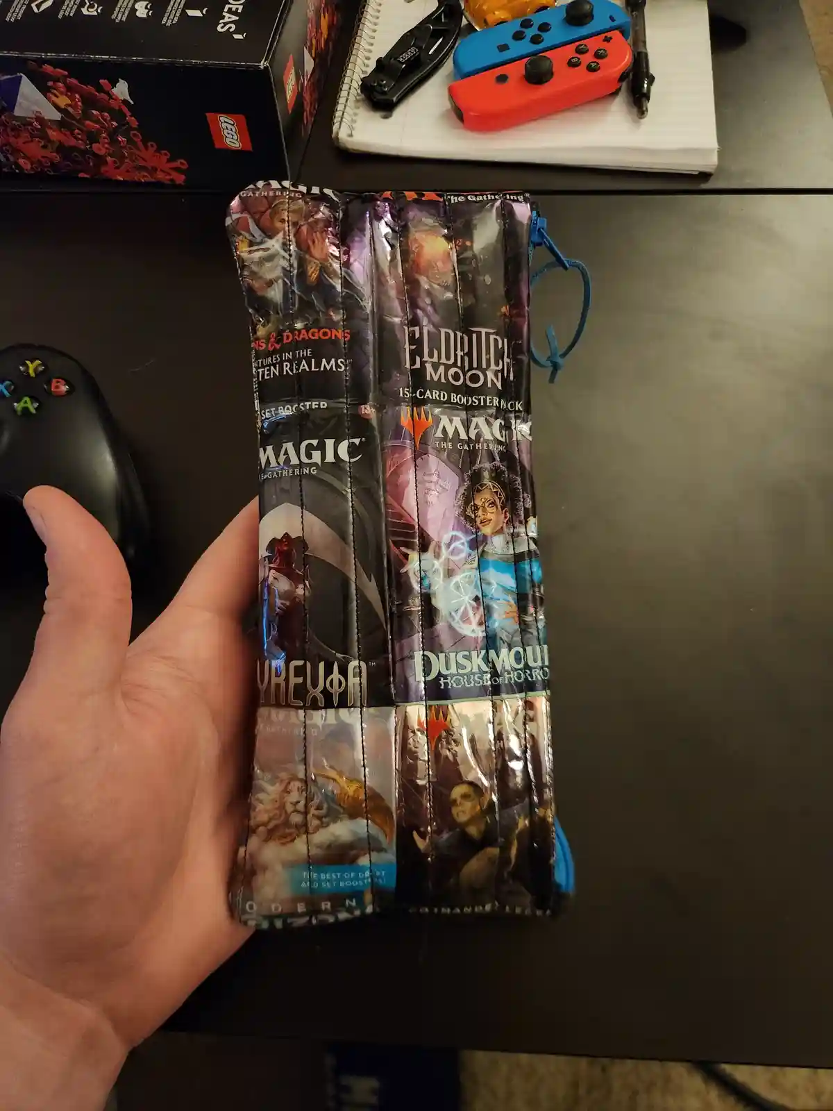 A hand holds a custom-made face mask crafted from multiple Magic: The Gathering booster pack wrappers, featuring sets like…