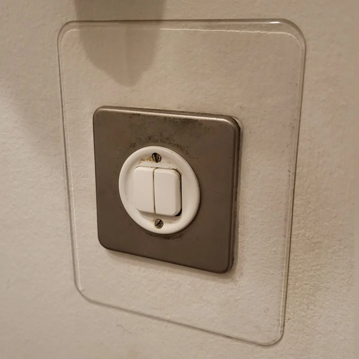 A close-up photo shows a white, dual-toggle light switch set in a tarnished silver metal plate, which is covered by a clear…