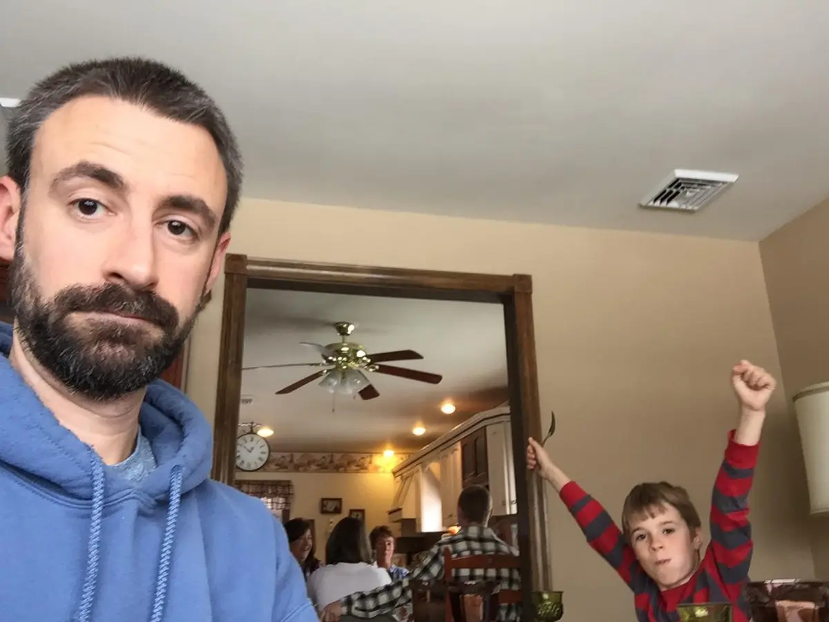 A man in a blue hoodie takes a selfie with a stern, unamused expression, while a young boy in a red and gray striped shirt…