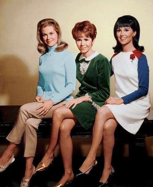 Three women in 1960s-style clothing sit side-by-side on a bench. The woman on the left wears a light blue turtleneck and…
