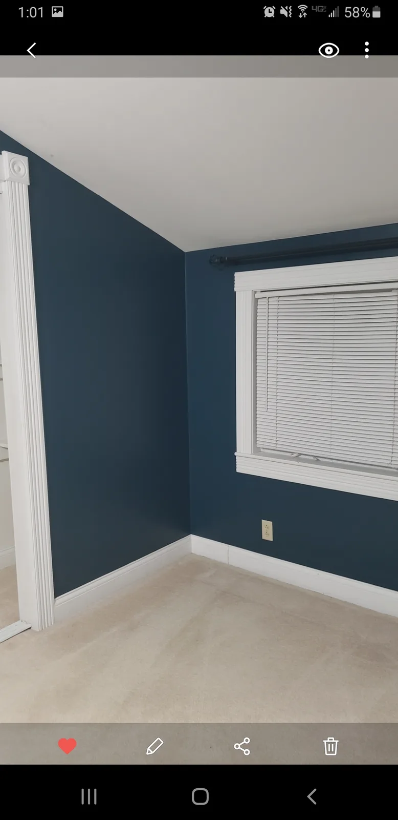 An empty room with dark blue walls, white trim, and beige carpet, featuring a window with closed blinds. The overlaid text…