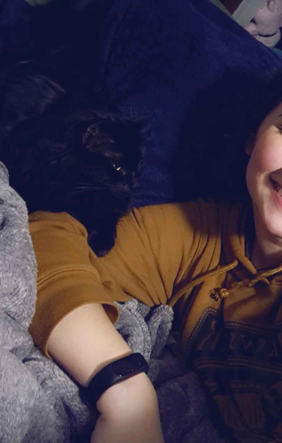A person in a mustard-yellow hoodie lies in bed, smiling, with a black cat curled up on their chest. The cat's glowing eyes…