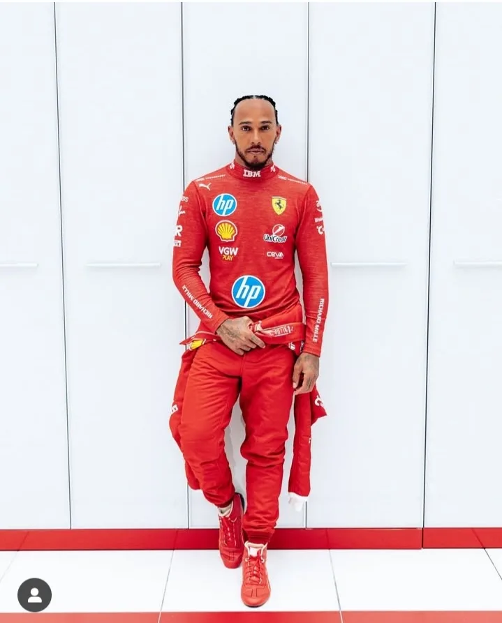a man in a full red Ferrari racing suit with sponsor logos like HP, Shell, and IBM stands against a white wall with red…