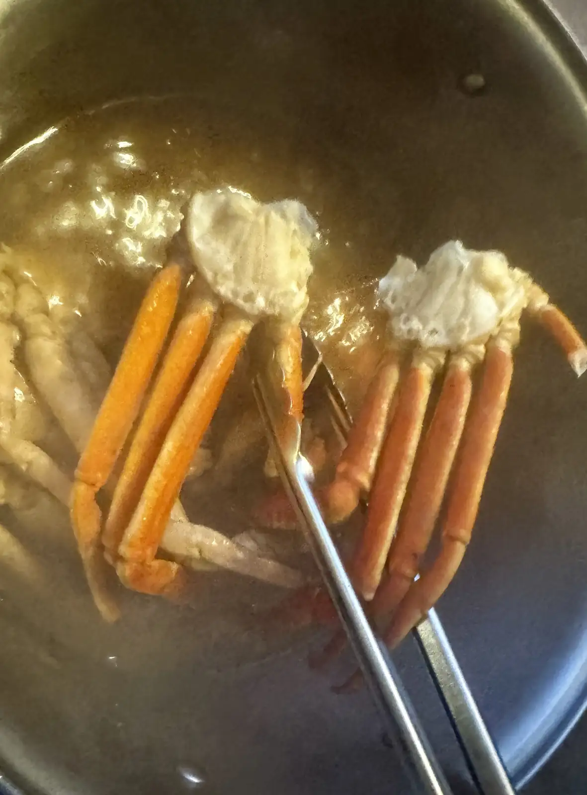 A close-up photo shows two cooked crab legs with orange shells and white meat being held by metal tongs over a pot of…