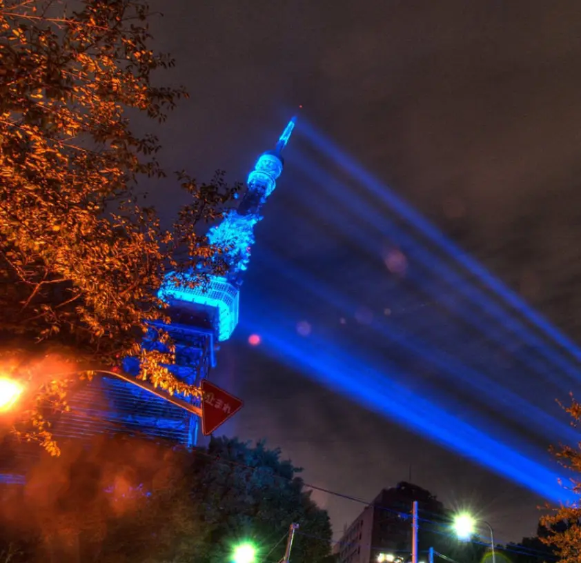 A nighttime photo of the Tokyo Tower illuminated in blue, with powerful blue spotlights projecting from its top. In the…