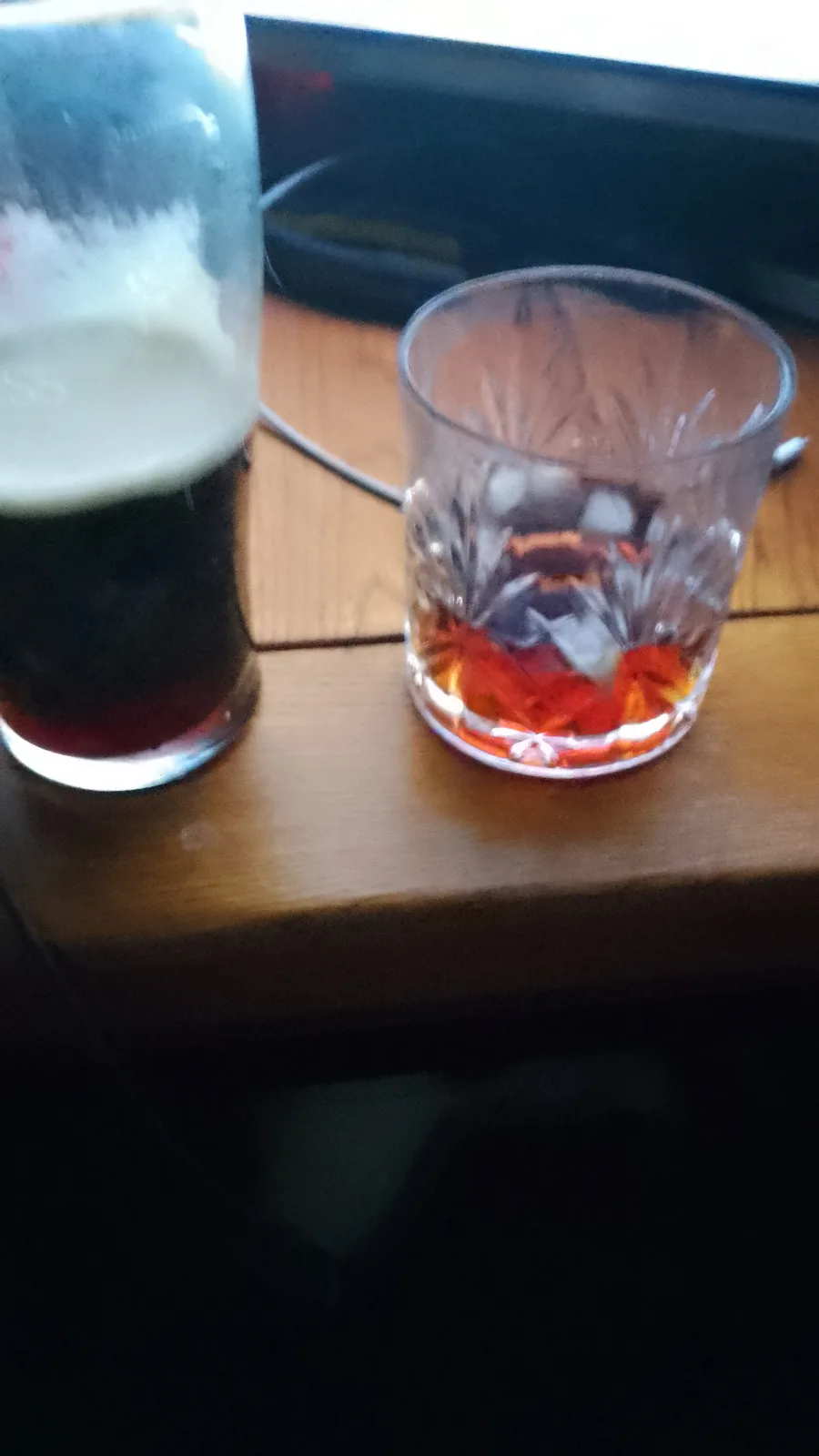 A close-up photo shows two drinks on a wooden surface: a tall glass of dark beer with a white head next to a short…