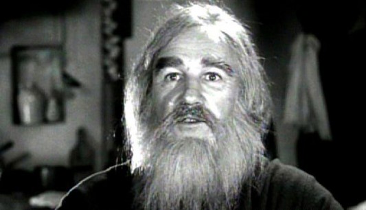 A black-and-white image shows a man with long white hair and a full beard, looking upward with a surprised or skeptical…
