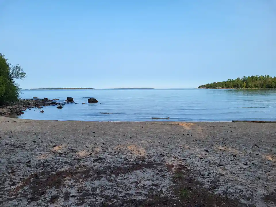 A serene beach scene with calm blue water, a sandy shore, and distant tree-covered islands under a clear sky. Overlaid text…