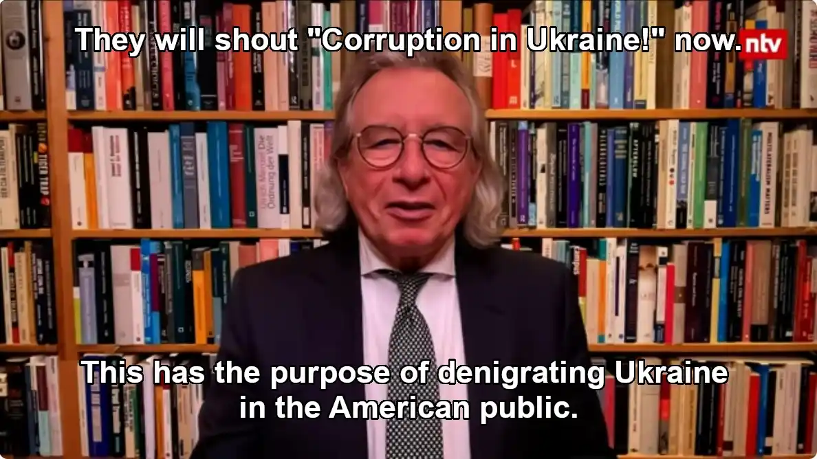 A man with glasses and long hair, wearing a suit and tie, is speaking in front of a bookshelf filled with books. White text…
