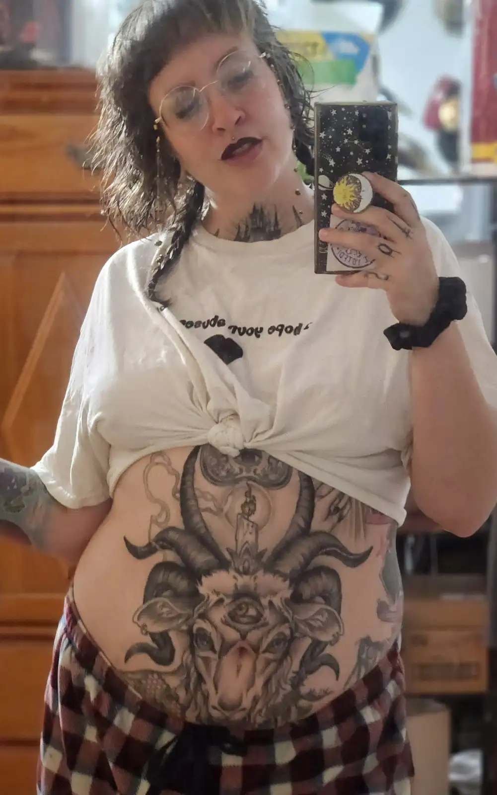 A person with dark hair and glasses takes a mirror selfie, holding a phone with a starry case. Their white t-shirt is tied…