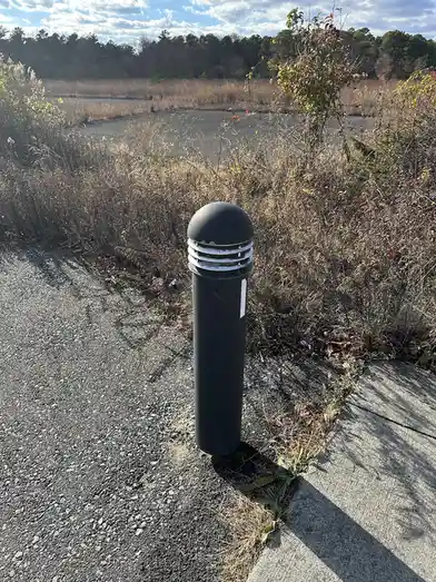 A black, cylindrical bollard with a domed top and white horizontal bands stands on a gravel path next to a concrete…