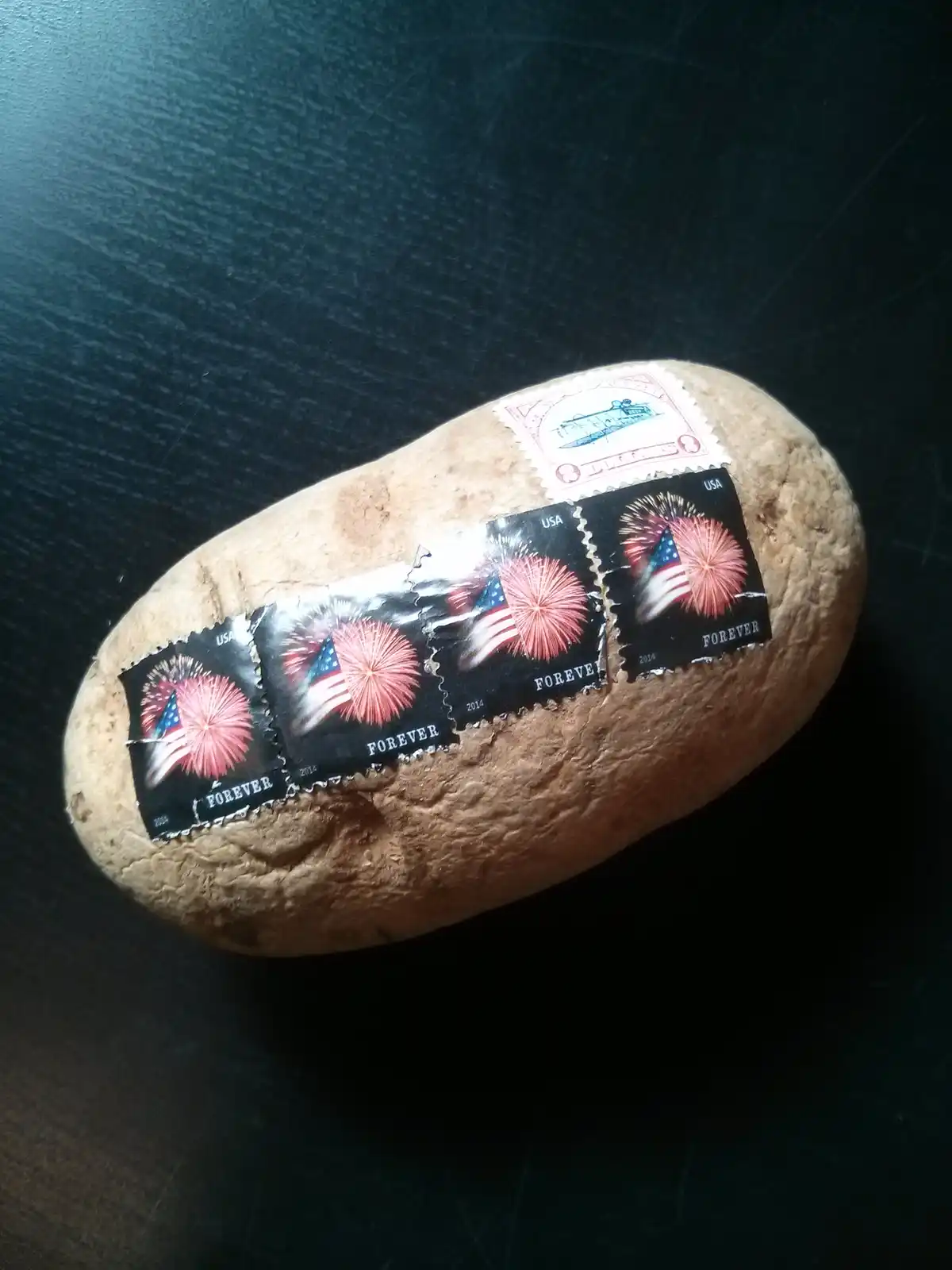 A potato sits on a dark surface with several U.S. postage stamps stuck to its skin, including four 'Forever' stamps…
