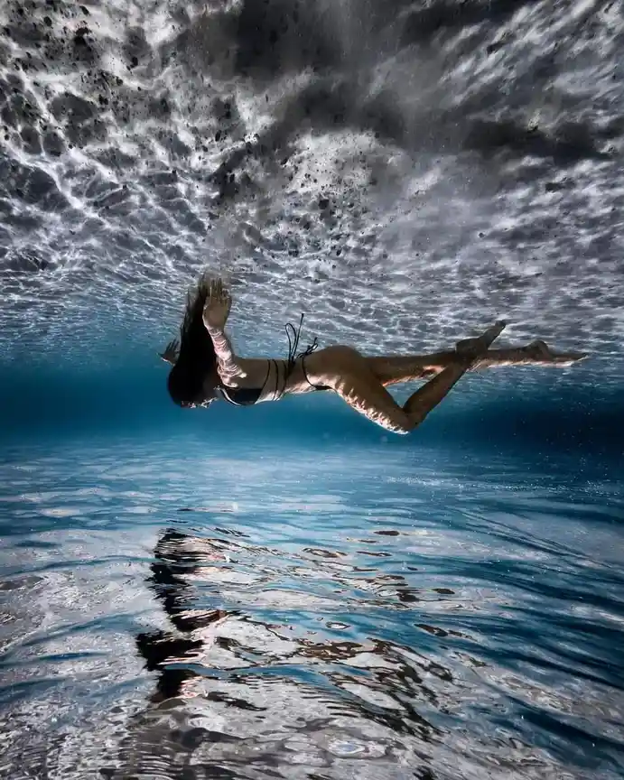 An underwater photograph shows a woman in a black bikini floating gracefully beneath the water's surface, her body angled…