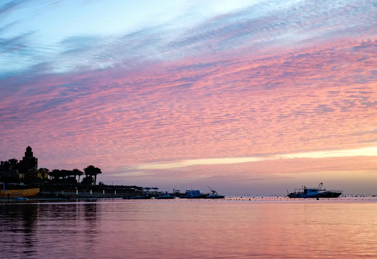 A serene sunset over a calm body of water with boats and a shoreline featuring palm trees and a building. The sky is painted…