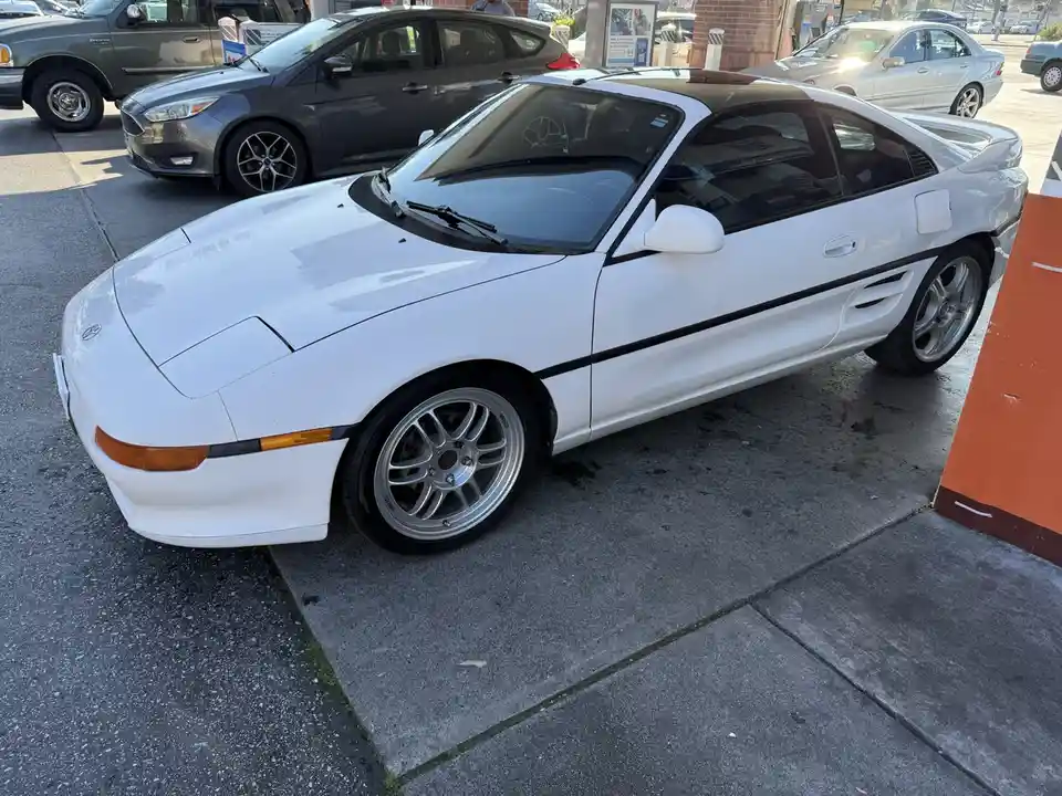 A white Toyota MR2 sports car is parked on a sidewalk next to a street, with other cars visible in the background. The car…