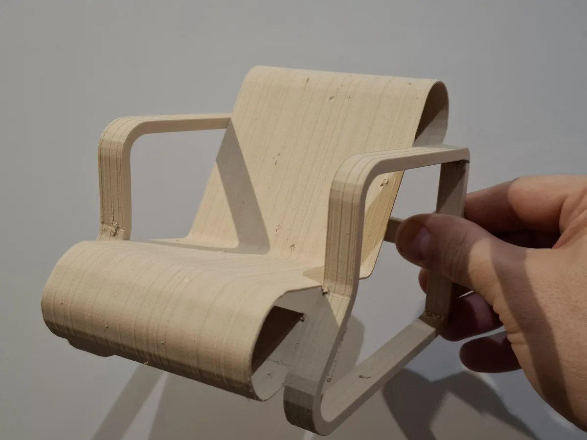 A hand holds a small, 3D-printed wooden chair model with visible layer lines, against a plain background. The overlaid text…