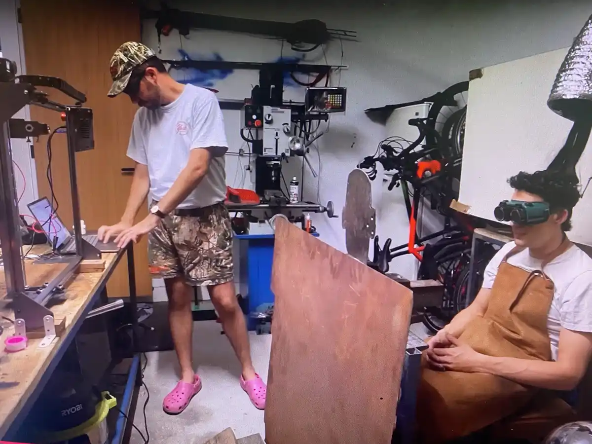In a cluttered workshop, one man in camouflage shorts and pink clogs stands at a 3D printer, while another man in an apron…