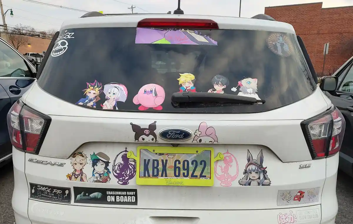 The rear of a white Ford Escape is covered in anime and pop culture stickers, including characters from games and shows…