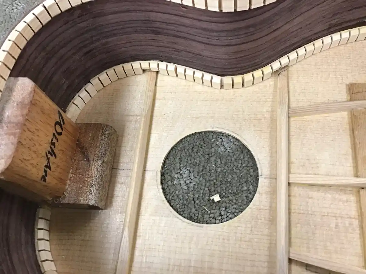 A close-up of a guitar body under construction, showing the soundhole surrounded by wood grain and a decorative binding. A…