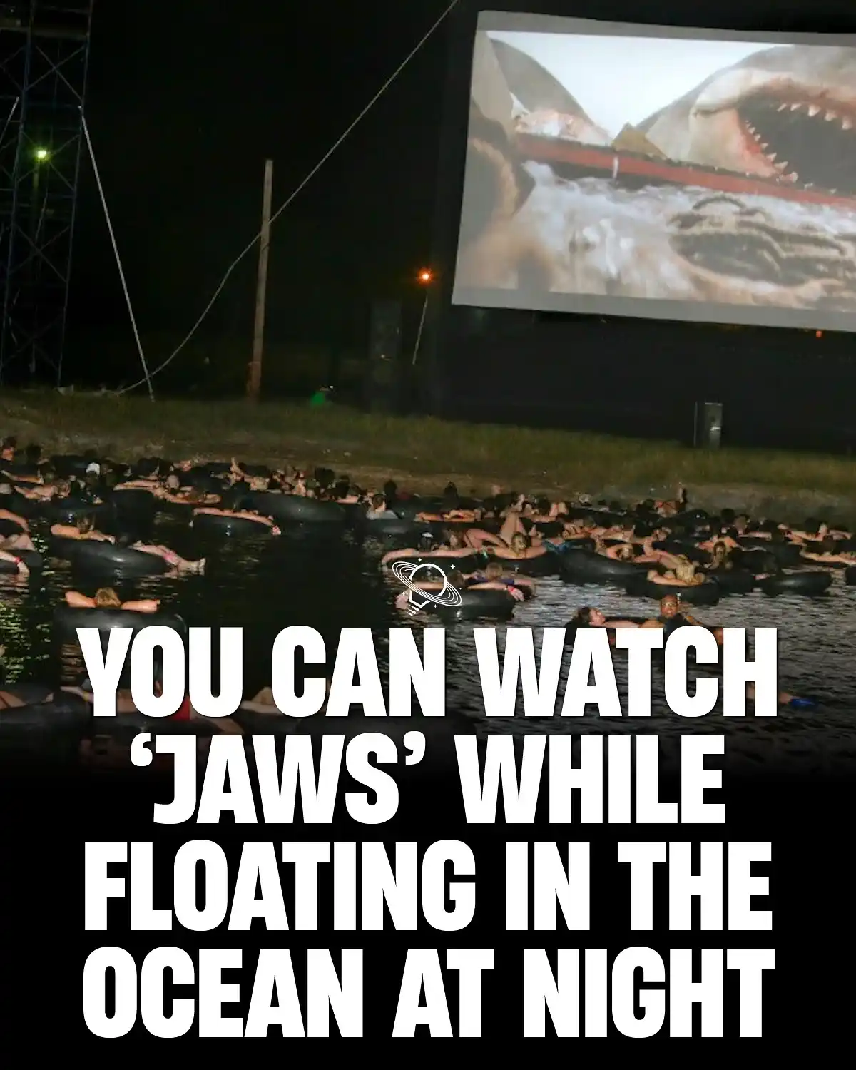 An outdoor movie screening at night shows a large screen displaying a scene from 'Jaws' with a shark attacking a boat…