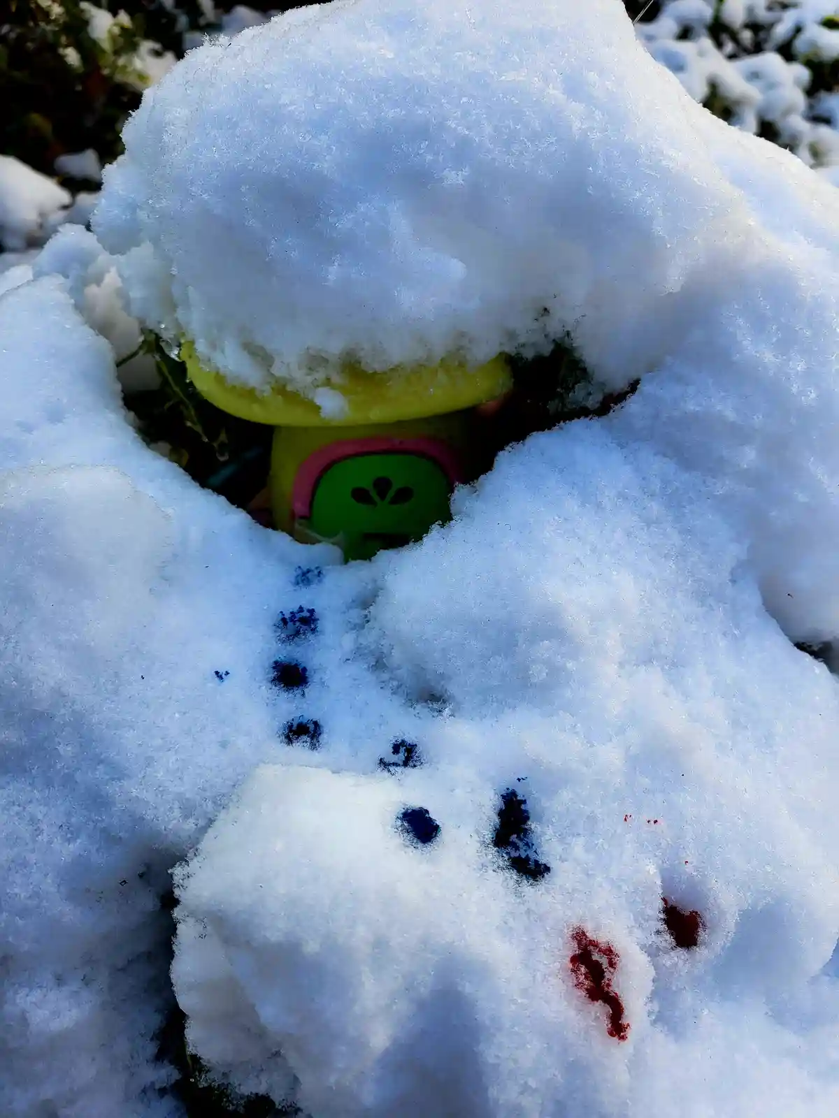 A colorful children's toy, partially buried under a thick layer of snow, has blue and red smudges on the snow in front of…