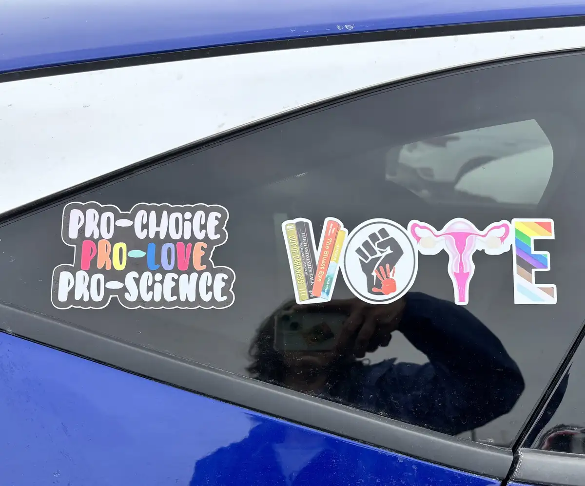A close-up of a blue car's rear window displaying several political and social justice stickers. On the left, a sticker…