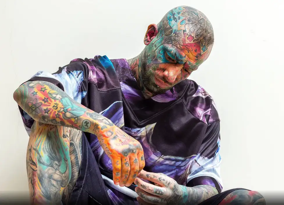 a man with extensive, colorful tattoos covering his entire face, head, arms, and hands, sitting and looking down while…