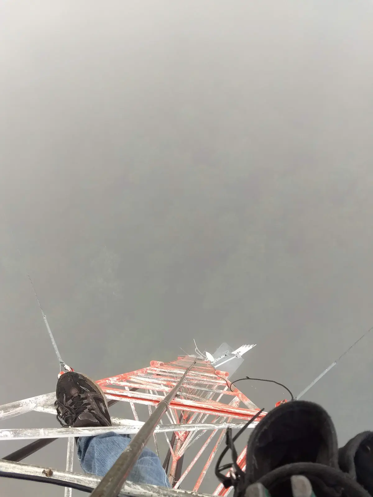 A first-person view from the top of a tall, red and silver metal communications tower, looking down through thick fog. The…