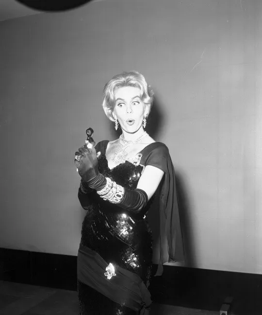A black-and-white photo of a glamorous woman in a sequined dress and gloves, holding a small object and making a surprised…