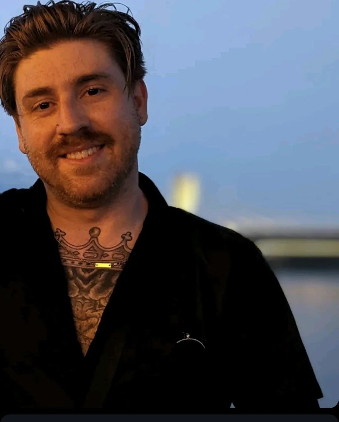 a man with a beard and short hair smiles at the camera while wearing a black jacket; a detailed crown tattoo is visible on…