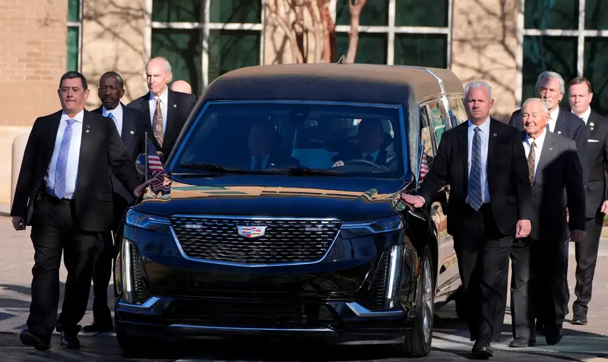 a black cadillac hearse with american flags on the side is parked outdoors, surrounded by men in dark suits. the vehicle has…